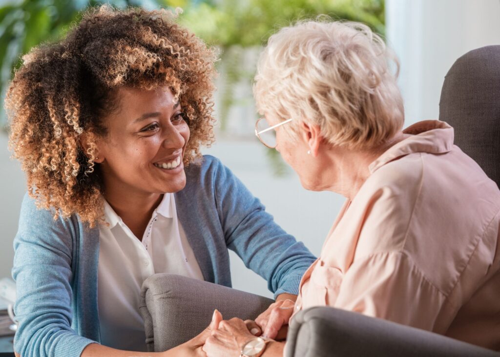 Younger African American woman in blue sweater holds hands of older woman in pink sweater.